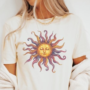 May include: A white t-shirt with a colorful sun graphic. The sun has a face with closed eyes and a smile. The sun's rays are purple, orange, and yellow.