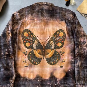 Puede incluir: Una camisa de franela de manga larga con un diseño de mariposa en la espalda. La camisa presenta un patrón de cuadros en tonos marrones y negros, con un efecto decolorado. La mariposa está detallada con acentos naranjas, amarillos y negros.