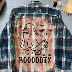 May include: A long-sleeved, button-down flannel shirt with a plaid pattern in shades of green, blue, and white. The back features a graphic of a duck dressed as a ghost, with the text "OOPS ALL BOOOOOTY" below. The shirt has a distressed, faded look.