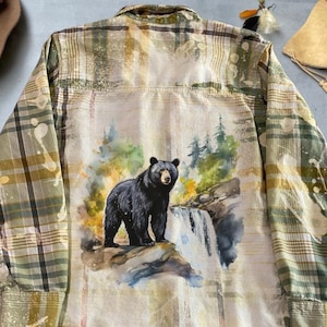 May include: A green and brown plaid flannel shirt with a watercolor painting of a black bear in a forest setting on the back.