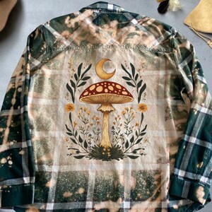 May include: A green and white plaid flannel shirt with a bleached effect. The back of the shirt features a mushroom with a crescent moon above it, surrounded by flowers and leaves.