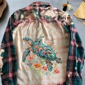 May include: A green and pink plaid flannel shirt with a bleached effect. The shirt features a graphic of a sea turtle with flowers on the back.