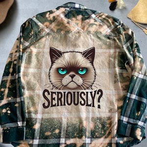 Könnte beinhalten: Ein grün-braunes kariertes Flanellhemd mit Bleich-Effekt. Das Hemd zeigt ein Cartoon-Bild eines mürrischen Katers mit dem darunter gedruckten Text "SERIOUSLY?"