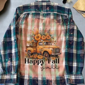 May include: A green and pink checked flannel shirt with a bleached effect. The shirt features a graphic of a vintage truck carrying pumpkins and flowers. The text "Happy Fall Y'all" is printed below the truck.