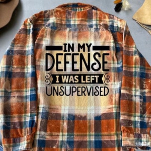 May include: A brown and blue plaid flannel shirt with the text "In my defense I was left unsupervised" printed on the back.