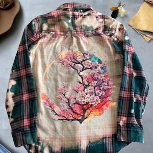Könnte beinhalten: Ein kariertes Flanellhemd in Grün, Rosa und Creme mit einem Blumendesign auf dem Rücken. Das Blumendesign zeigt einen Baum mit rosa Blüten und einer goldenen Halbmondform. Das Hemd hat lange Ärmel und eine Knopfleiste.