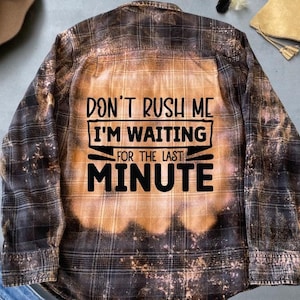 Może przedstawiać: Koszula flanelowa w czarno-brązową kratę z wybielonym wzorem. Na plecach koszuli znajduje się napis "Don't rush me I'm waiting for the last minute".