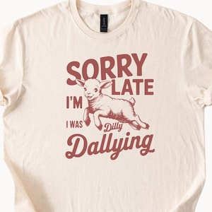 May include: Cream-colored t-shirt with the text "SORRY I'M LATE I WAS Dilly Dallying" in maroon. A cartoon lamb is illustrated in the center, also in maroon. The shirt has short sleeves and a crew neck.