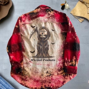 May include: Red and black checked flannel shirt with a bleached design featuring a cartoon grim reaper with a scythe and the text "It's Got Pockets".
