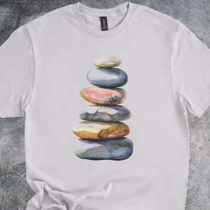 May include: White t-shirt featuring a watercolor illustration of a stack of balanced stones. The stones are in shades of gray, orange, and yellow. The shirt has a crew neck and short sleeves.