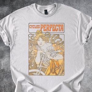 May include: White t-shirt with an Art Nouveau design of a woman with flowing hair and a bicycle. The text "CYCLES PERFECTA" is in an orange box above the illustration. The shirt has a crew neck and short sleeves. The background is grey.