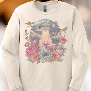 Puede incluir: Camiseta de manga larga color crema con un gráfico colorido. El diseño incluye un OVNI, pájaros y un arreglo floral con flores rosas, azules y naranjas. La camiseta tiene cuello redondo y puños acanalados.