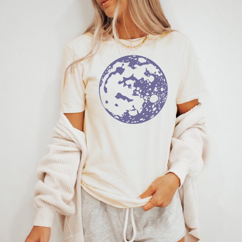 Op de afbeelding: Een wit T-shirt met een paarse maanprint. De maan is een gestileerde illustratie met een witte binnenkant en een paarse omtrek.