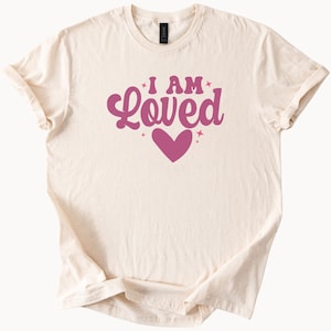 Puede incluir: Camiseta color crema con las palabras "I AM Loved" en una fuente de estilo retro, con un gr&aacute;fico de coraz&oacute;n. El texto y el coraz&oacute;n son de color rosa intenso. La camiseta tiene mangas cortas y cuello redondo.