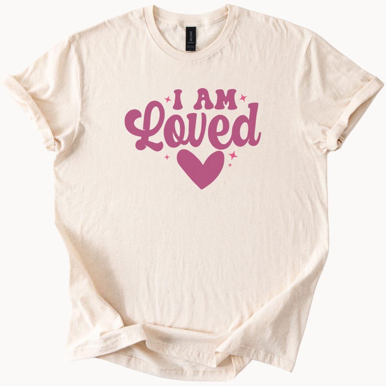 Puede incluir: Camiseta color crema con las palabras "I AM Loved" en una fuente de estilo retro, con un gr&aacute;fico de coraz&oacute;n. El texto y el coraz&oacute;n son de color rosa intenso. La camiseta tiene mangas cortas y cuello redondo.