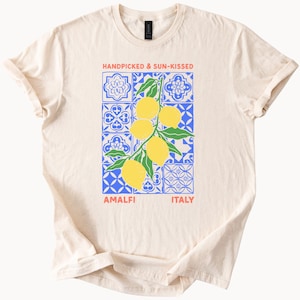 May include: Cream-colored t-shirt featuring a graphic of lemons on a branch, set against a blue and white tile pattern. The text reads "HANDPICKED & SUN-KISSED" above and "AMALFI ITALY" below the graphic.