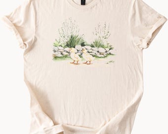 Puede incluir: Camiseta color crema con una ilustración en acuarela de dos patitos amarillos cerca de un muro de piedra y vegetación. El diseño utiliza una paleta de colores suave y natural, con los patos como punto focal. La camiseta tiene cuello redondo clásico y mangas cortas.