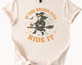 Könnte beinhalten: Cremefarbenes T-Shirt mit einer Grafik einer Hexe auf einem Besen, die eine Tasse hält. Der Text lautet "IF THE BROOM FITS RIDE IT" in Orange. Ein Fledermaus- und Stern-Design ist ebenfalls enthalten.