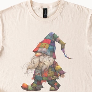 May include: Cream-colored t-shirt featuring a whimsical illustration of a gnome. The gnome is depicted with a long white beard, a large orange nose, and a patchwork hat and outfit in various colors. The gnome is walking.