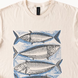 May include: Cream-colored t-shirt featuring a repeating pattern of stylized fish in shades of blue and gray. The fish are arranged in a block print style, with detailed scales and fins. The shirt has a crew neck and short sleeves.