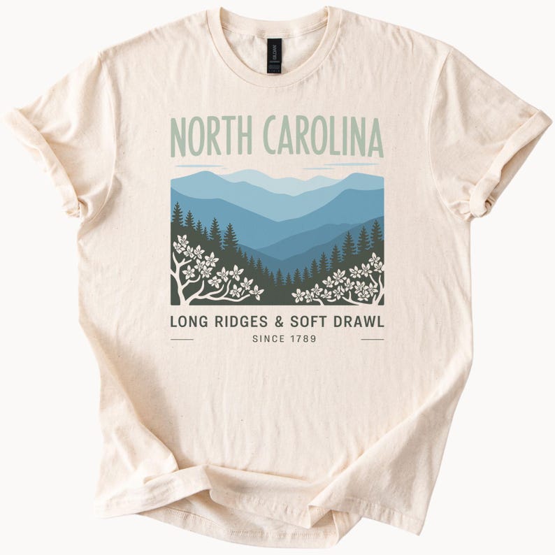 May include: Cream-colored t-shirt featuring a graphic of North Carolina mountains in shades of blue and green. The text "NORTH CAROLINA" is above the image, with "LONG RIDGES & SOFT DRAWL" and "SINCE 1789" below.