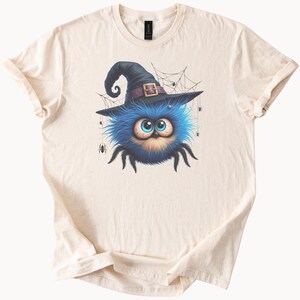 Pode incluir: T-shirt creme com um design de Halloween. O gráfico mostra uma criatura azul e peluda com olhos grandes, usando um chapéu de bruxa e cercada por aranhas e teias de aranha. O design está centrado na frente da camiseta.