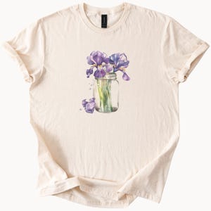 May include: Cream-colored t-shirt featuring a watercolor illustration of purple irises in a glass jar. The design includes a few loose petals and water droplets. The shirt is made of soft fabric and has a classic crew neck.