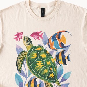 May include: A cream-coloured t-shirt with a vibrant sea-themed design. It features a green and yellow turtle, surrounded by colourful fish in shades of pink, orange, blue, and yellow, with blue and purple leaves.