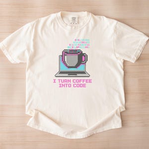 May include: An off-white short-sleeve t-shirt featuring a pixel art graphic of a laptop with a coffee mug on the screen. Above the mug is binary code in pink and blue. The text below the laptop reads, I TURN COFFEE INTO CODE in pink letters.