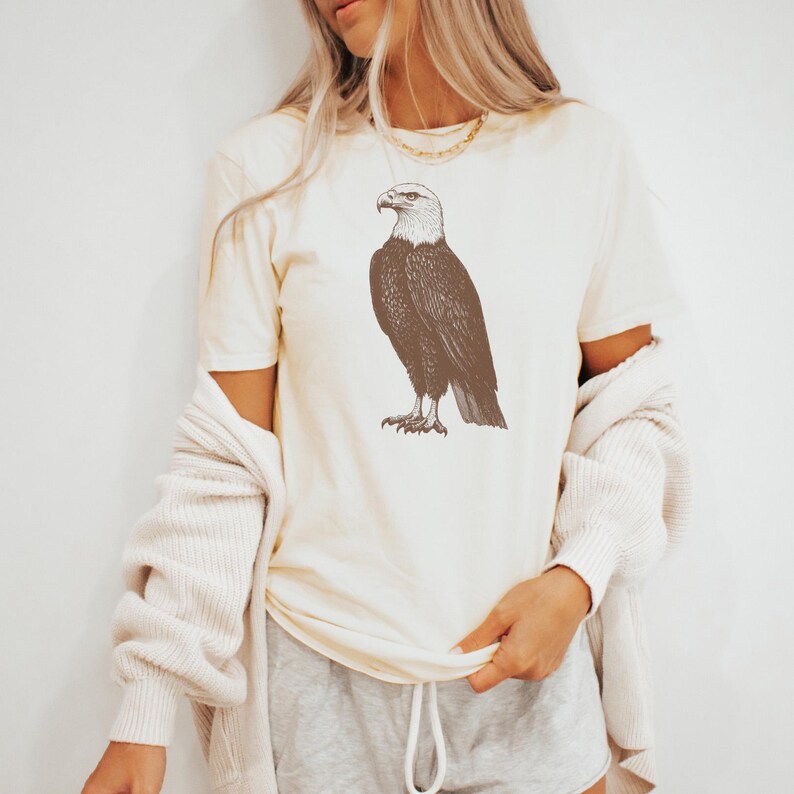 May include: A cream-colored t-shirt with a brown line drawing of a bald eagle on the front.