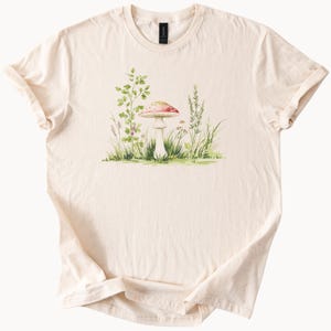 Könnte beinhalten: Cremefarbenes T-Shirt mit einer Aquarellillustration eines rotkappigen Pilzes mit weißen Punkten, umgeben von grünem Gras und Blättern. Das Design hat ein skurriles, von der Natur inspiriertes Thema.