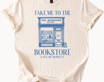 May include: Cream-colored t-shirt with a blue bookstore graphic. The graphic includes the text "TAKE ME TO THE BOOKSTORE & TELL ME I'M PRETTY". The bookstore design features the text "THE BOOKMINE EST.1983" and "BOOKS & POSTCARDS".