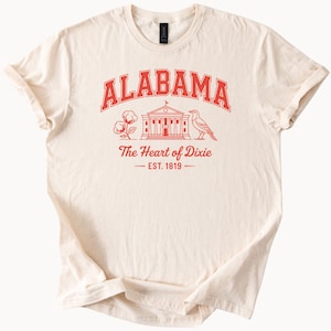 Pode incluir: Uma camiseta de cor creme com um design gráfico vermelho que diz "Alabama, The Heart of Dixie, Est. 1819". O gráfico também inclui um edifício, uma bola de algodão e um pássaro.