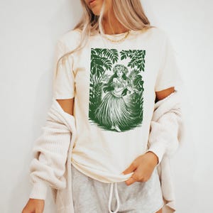 May include: A white t-shirt with a green print of a hula dancer in a tropical setting. The dancer is wearing a flower crown and a grass skirt.