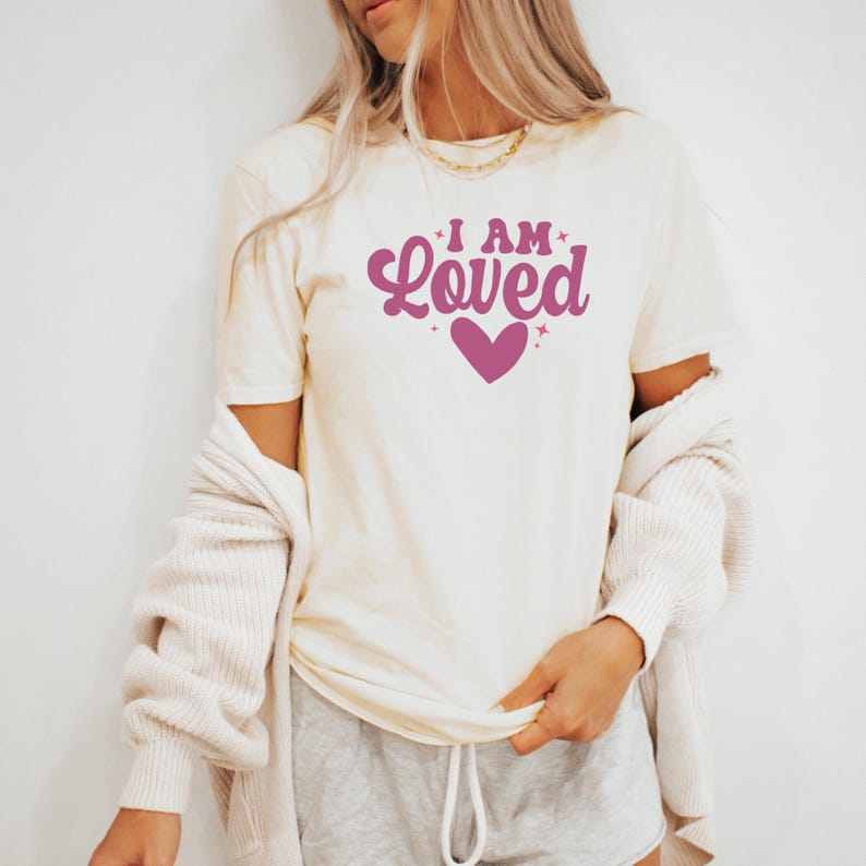 Puede incluir: Camiseta color crema con el texto "I AM Loved" en fuente cursiva, con un gr&aacute;fico de coraz&oacute;n. La camiseta se combina con pantalones cortos grises y una rebeca color crema.
