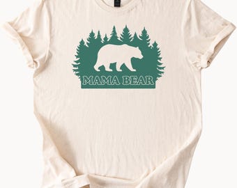 Puede incluir: Una camiseta de color crema con una silueta verde de un oso caminando por un bosque con el texto "Mama Bear" debajo.