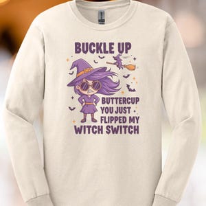 Op de afbeelding: Crèmekleurig shirt met lange mouwen met een afbeelding van een cartoon heks in een paarse hoed en jurk, met een zonnebril. De tekst luidt: "BUCKLE UP BUTTERCUP YOU JUST FLIPPED MY WITCH SWITCH."