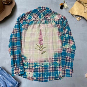 May include: A long-sleeved plaid shirt in teal, pink, and yellow, with a bleached center featuring a pink floral design. The shirt has a button-down front and cuffs, and a classic collar.