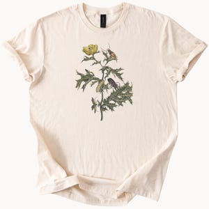 May include: A cream-colored t-shirt with a vintage illustration of a yellow flower with green leaves and various insects.
