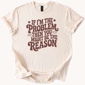May include: Cream-colored t-shirt with the phrase "If I'm the problem then you might be the reason" in brown lettering. The text is in a retro, groovy font. The shirt is made of a soft material and has rolled-up sleeves.