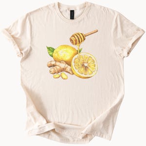 May include: A white t-shirt featuring a watercolour illustration of a lemon, ginger, and honey. The lemon is sliced in half with honey dripping from a honey dipper.