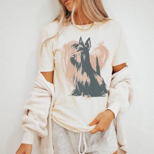 May include: Cream-colored t-shirt featuring a stylized illustration of a Scottish Terrier dog within a pink heart shape. The dog is depicted in shades of gray and black. The shirt has short sleeves and a crew neck.