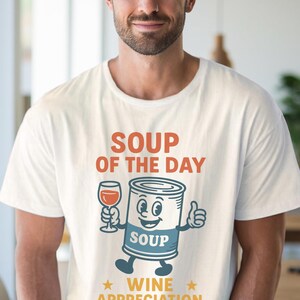 Op de afbeelding: Wit T-shirt met de tekst "SOUP OF THE DAY" in oranje. Een cartoon soepblik karakter houdt een wijnglas vast en geeft een duim omhoog. De tekst "WINE APPRECIATION" staat eronder.