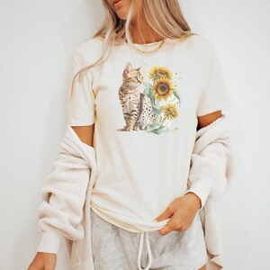 May include: A cream-coloured t-shirt featuring a graphic of a brown tabby cat sitting in a field of sunflowers. The cat is looking to the right and the sunflowers are in bloom.