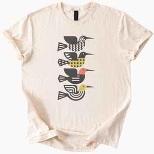 May include: A cream-colored short-sleeve t-shirt featuring a vertical graphic of four stylized birds stacked on top of each other. Each bird has a unique geometric pattern in black, yellow, and coral, including zigzags, dots, and circular lines.