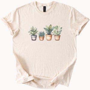 May include: Cream-coloured t-shirt with a watercolour illustration of four potted plants. The plants vary in size and type, with green and grey foliage. The pots are in shades of grey and tan.
