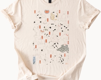 May include: Cream-colored t-shirt featuring a Japanese-inspired illustration of various dogs in black and white. The design includes red and orange accents and Japanese text. The shirt has a crew neck and short sleeves.