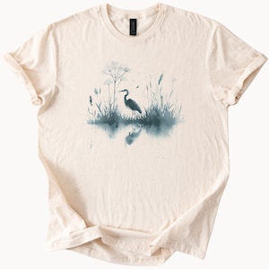 May include: A beige t-shirt with a watercolor design of a heron standing in a marsh with tall grasses and a reflection in the water.