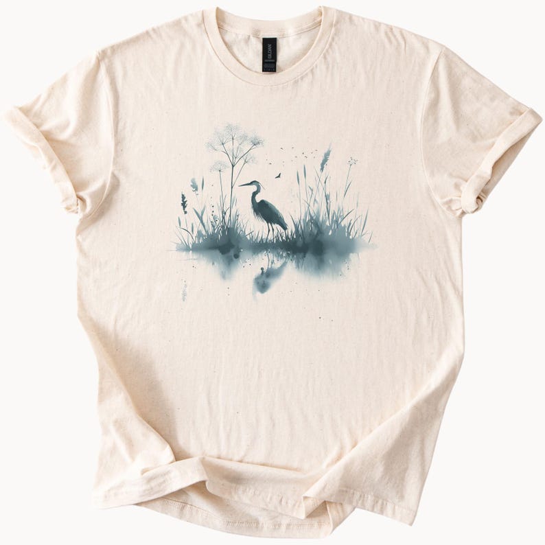 May include: A beige t-shirt with a watercolor design of a heron standing in a marsh with tall grasses and a reflection in the water.