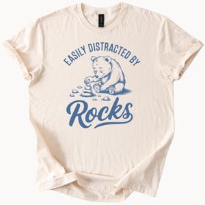 May include: A beige t-shirt with a blue graphic of a bear stacking rocks. The text "Easily Distracted By Rocks" is printed on the shirt.